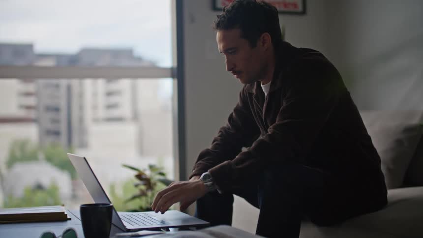A man sitting in front of his laptop working on something and then pausing to pick up his pen and deep in thought