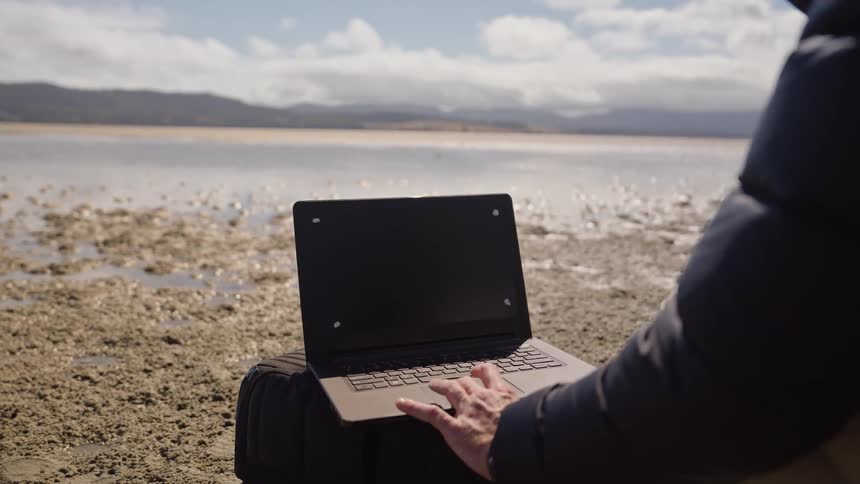 A person types on his laptop by the water
