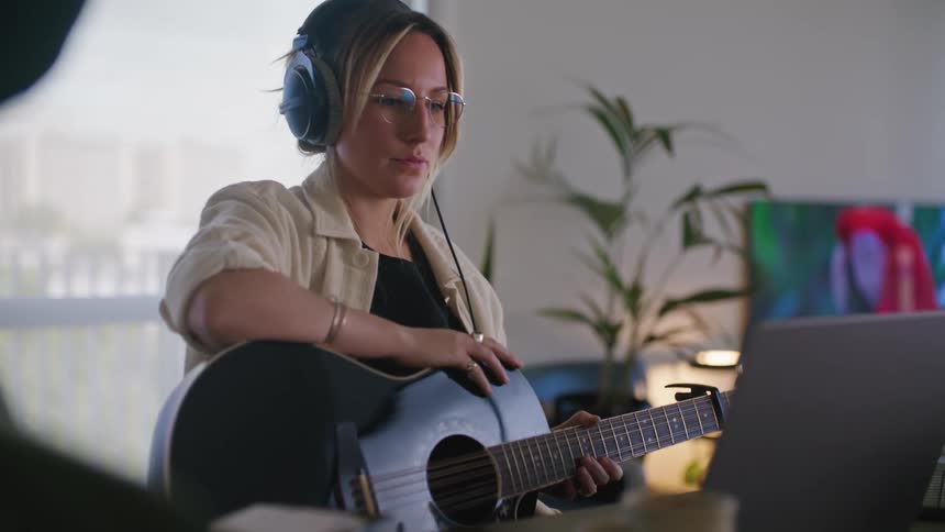 Woman holding acoustic guitar nods and taps to music she is listening to on headphones
