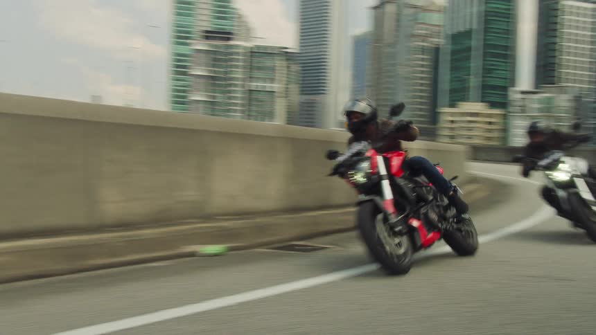 Two motorcycles race around a curve with tall buildings and skyscrapers in the distance