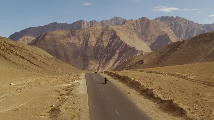 Motorcyclist follows other vehicles along road through desert passing toward mountains on a lightly cloudy day