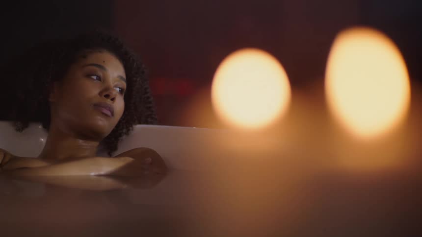 A somber beautiful black woman is soaking in a bathtub by candlelight