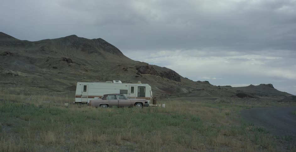 A car and rv park on a grass plain before a hill