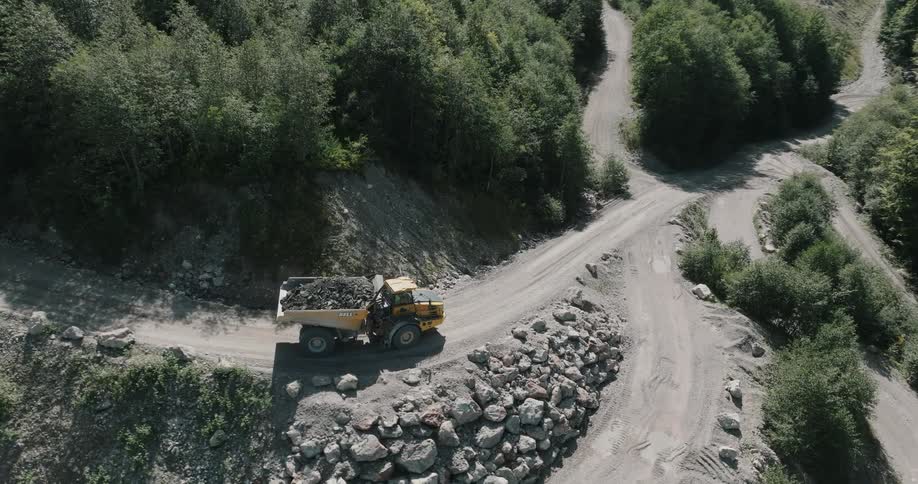 A yellow dump truck filled with gravel driving down a gravel road near a forest
