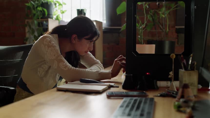 A woman is sitting in an office at a large desk adjusting the dial on a clear box with a black frame that is on the desk beside a computer monitor and keyboard in a room with plants and the sun shinning through the window built into the red brick wall