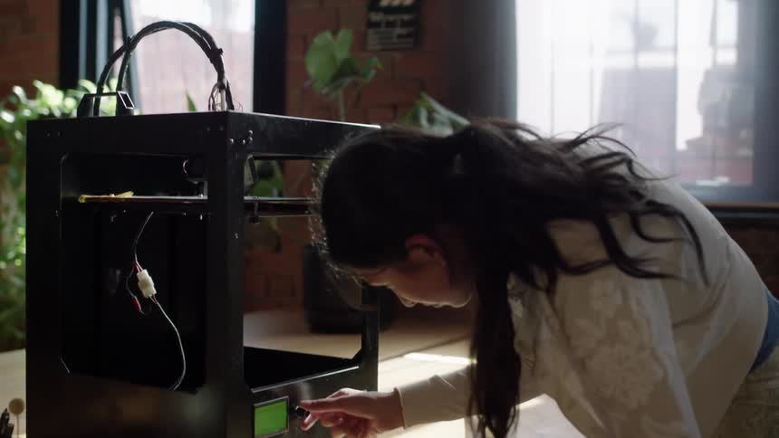 A woman is operating a 3d printing machine as she turns a knob and stands up to look inside
