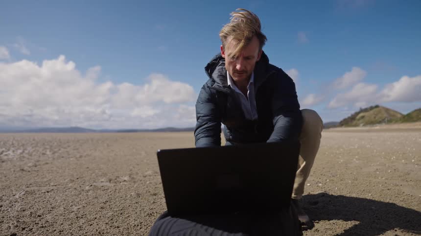 A man crouching in front of a laptop on the sandy ground among a desert landing