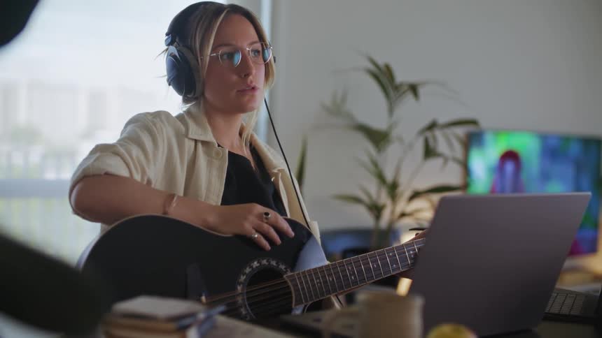 A woman in her living room is wearing headphones and using a laptop to help her create music with her acoustic guitar