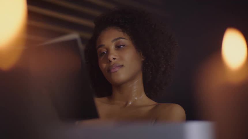 A beautiful black woman is reading a book while soaking in a bathtub under candlelight