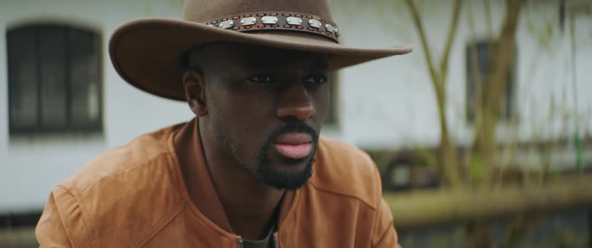 A man walks in front of a house as he puts a cowboy hat on his head and he stops and leans forward as he takes a drink from a bottle