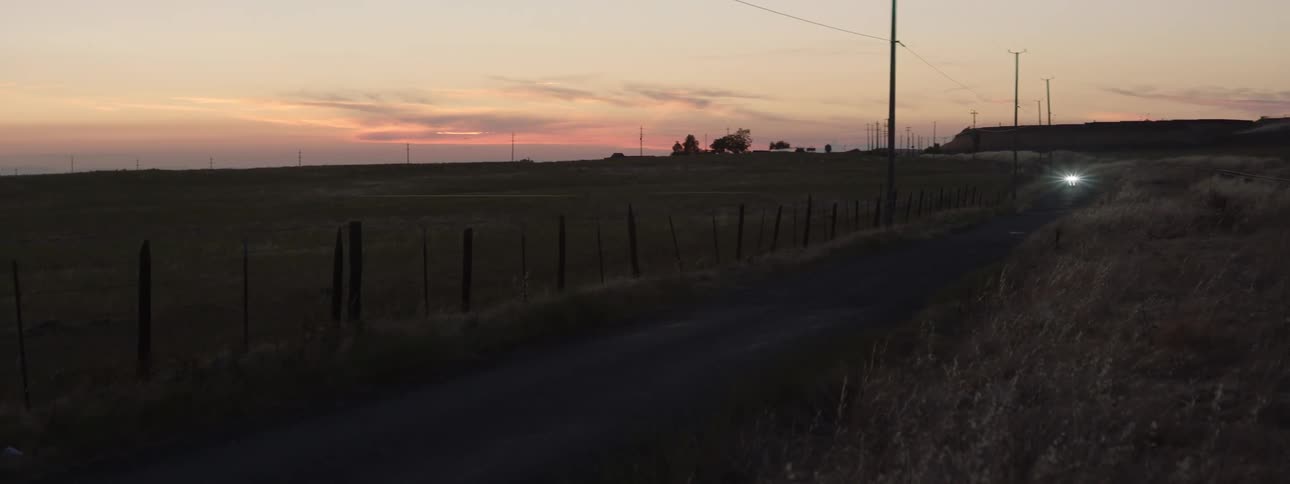 A motorcycle with the headlight on travels on a rural road as the sun has set