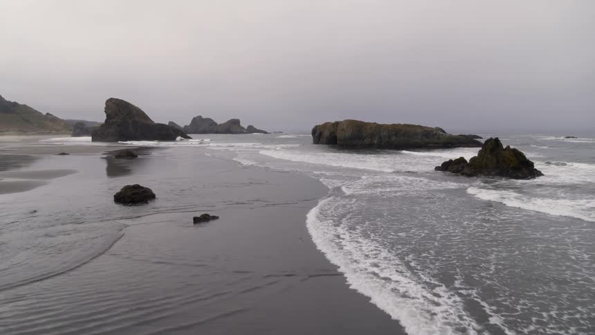 Gentle waves wash ashore around large rocks and sea stacks on a glassy smooth beach