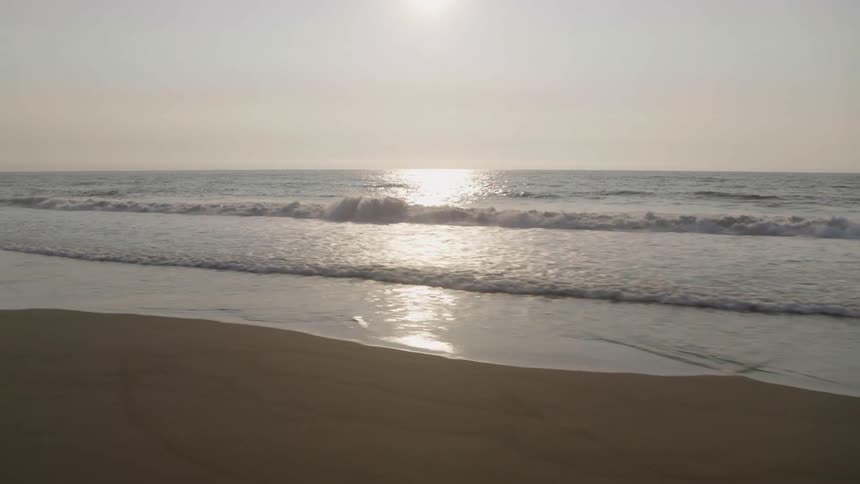 Low traverse over the perfectly smooth sand of a secluded beach as sunshine reflects from gentle breaking waves rolling in from open ocean
