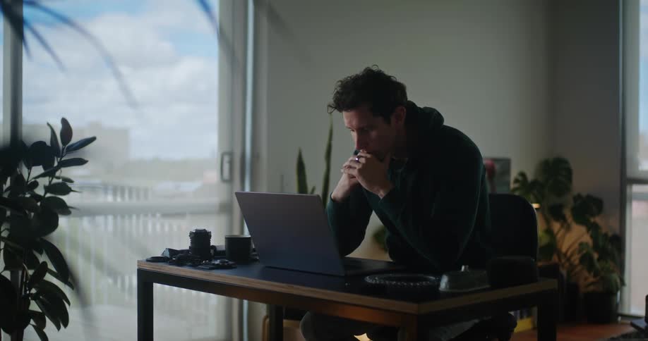 A man sitting at his desk picking up camera equipment and then using his laptop in front of him