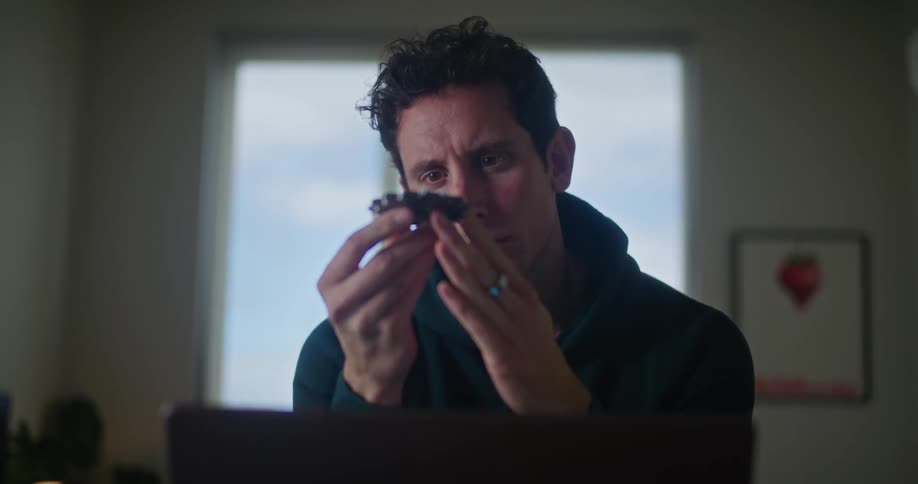 A man is sitting at a desk in front of a laptop computer as he looks at a round wheel gear in his hands and he gets up and leaves