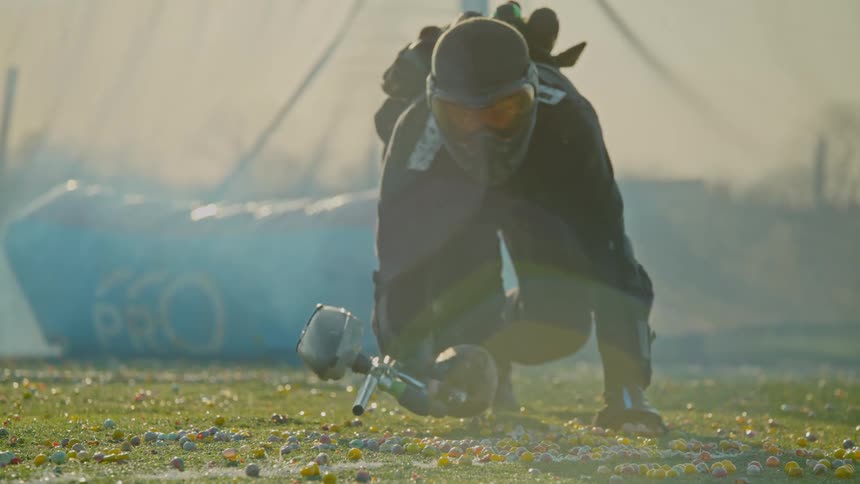 A paintball player in full gear is falling to the ground with his paintball hand in hand and landing on a turf filled with paintballs and a blowup barricade behind him