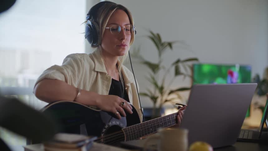 A woman sits with her acoustic guitar in front of her laptop while listening to music