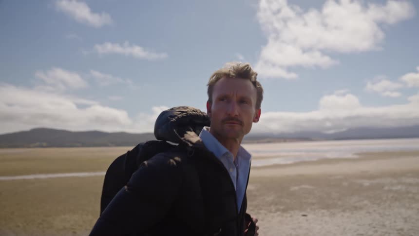 A man wearing a jacket and a backpack is walking across the beach and there are mountains in the distance under a cloudy sky