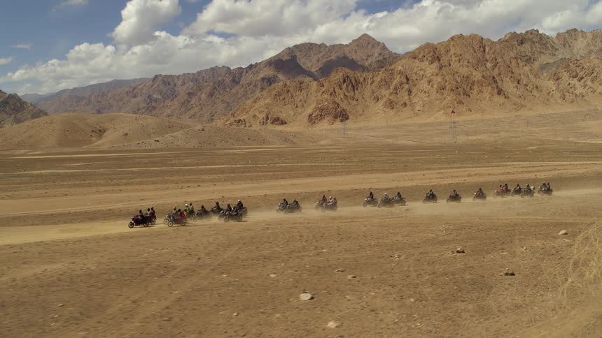 A group of around 20 motorcycle riders are riding bikes down a dirt path in the middle of a dusty desert with large dirt hills behind them