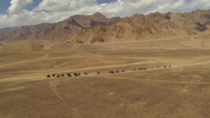 A caravan of motorcycles drive along a dusty dirt trail over a brown canyon in the day