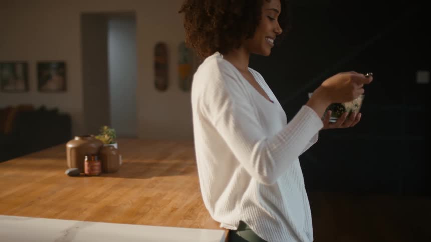 A woman standing in her kitchen eating food out of a bowl while her dog stands next to her looking for attention