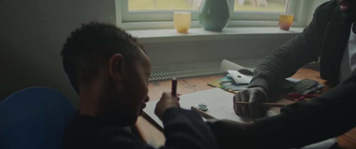 Father sitting with his son on a table next to a window at home helps him with homework