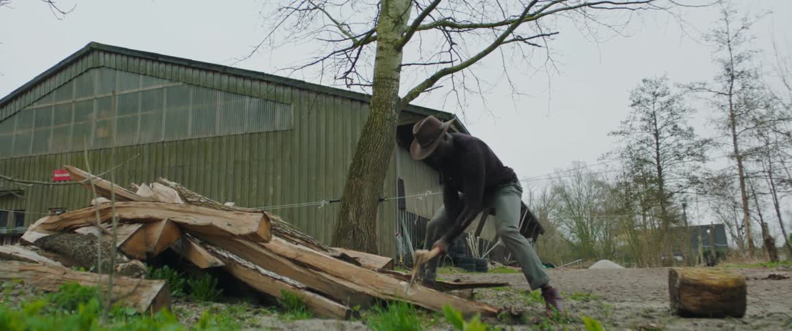 A black man is vigorously chopping wood in front of a large green barn