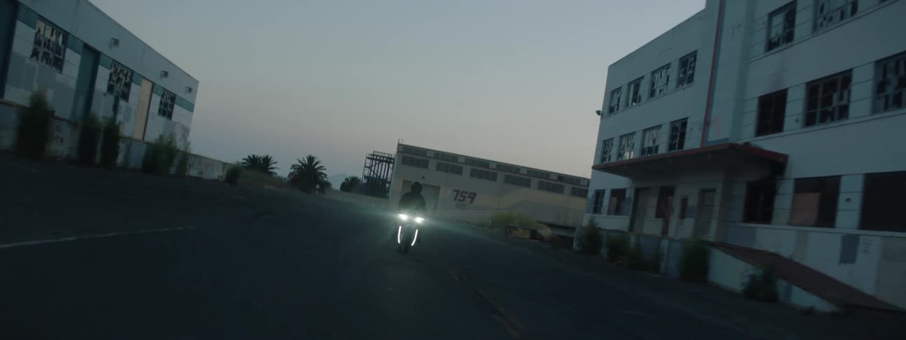 A person wearing all black riding a motorcycle on a road in between buildings at dusk