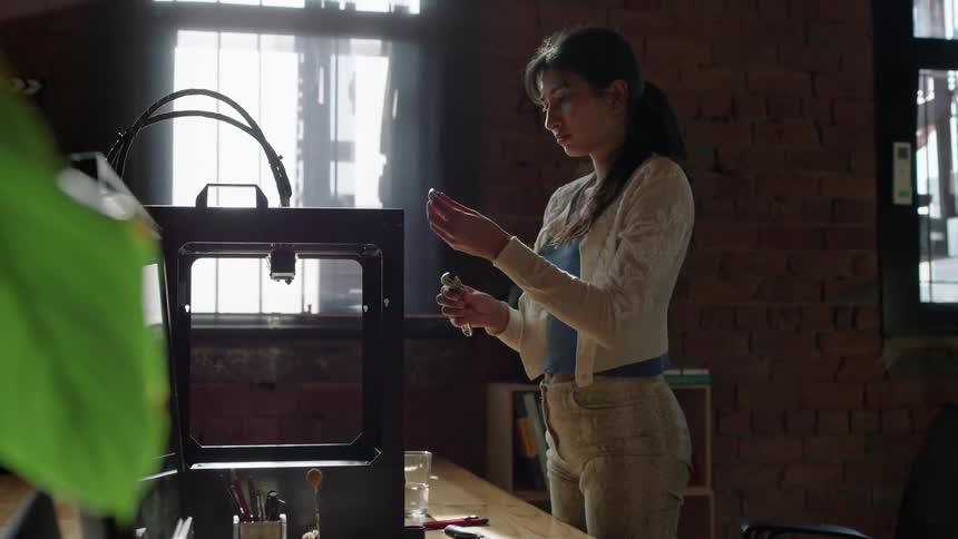 A woman standing in front of a mechanical device on her desk next to a laptop while she leans over and looks at the device