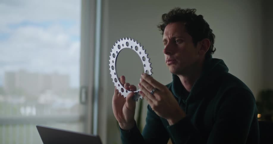 A man is sitting at a desk in front of a laptop computer as he looks at a round wheel gear and types on the keyboard