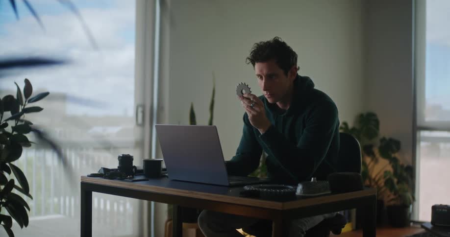 Man with hoodie works on laptop holding up pinion in room