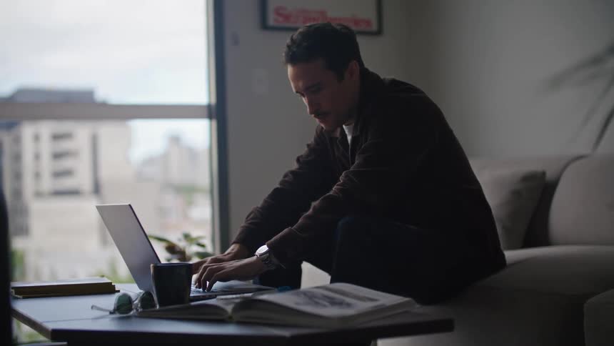 A man sitting on his couch looking at a book while typing on his laptop in front of him