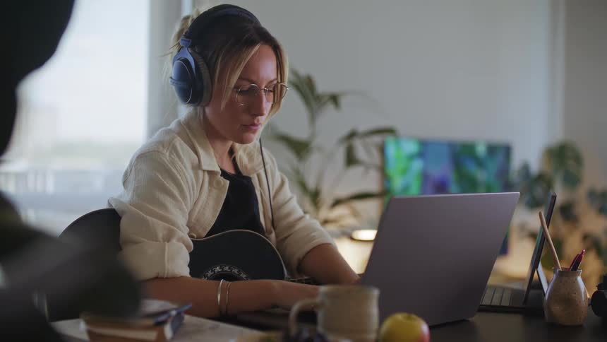 A woman sitting at a desk wearing headphones with an acoustic guitar in her lap is working on a laptop computer