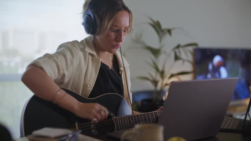 Woman wearing headphones records herself playing acoustic guitar in front of two laptops