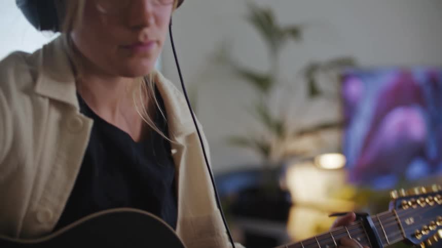 A woman is playing a song on guitar listening to something on headphones