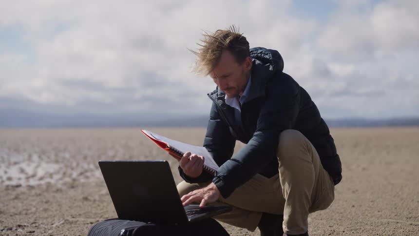 A man kneeling in front of his laptop sitting on a bag on the ground as he holds a notebook in the desert