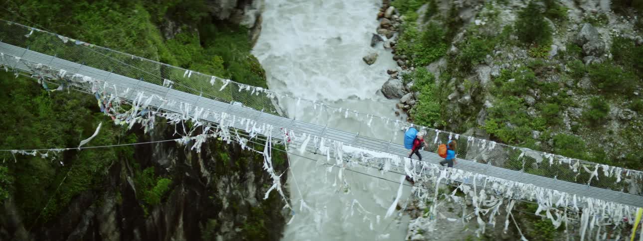 Two hikers walk over a suspension bridge that goes over a rushing river in a ravine