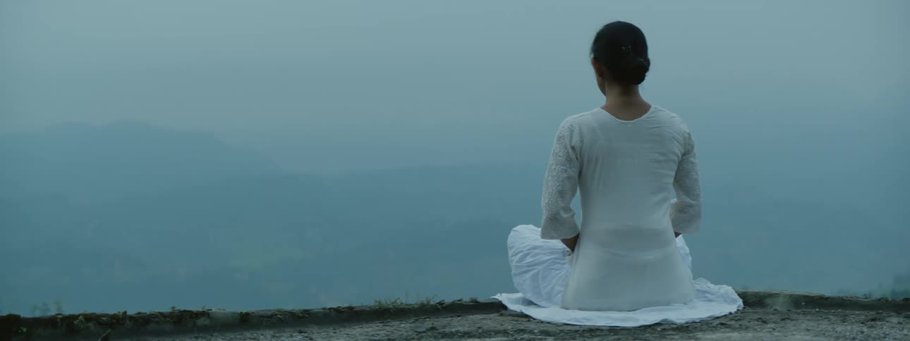 A woman in a white gown sits and meditates cliffside near hazy hills and valleys