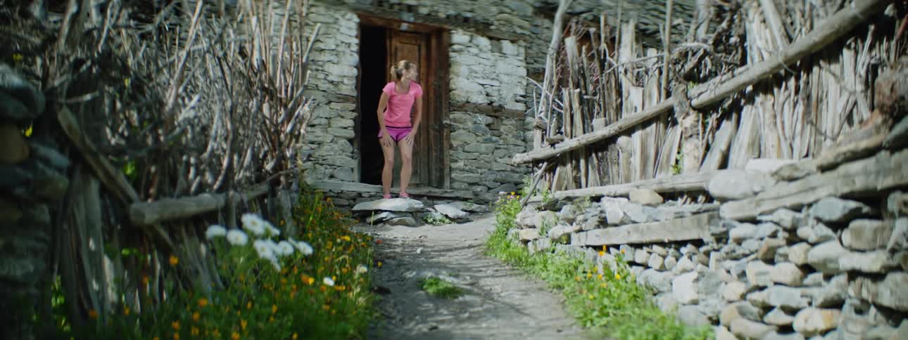 Woman with ponytail pink shirt and shorts puts on sneakers in front of front door of building