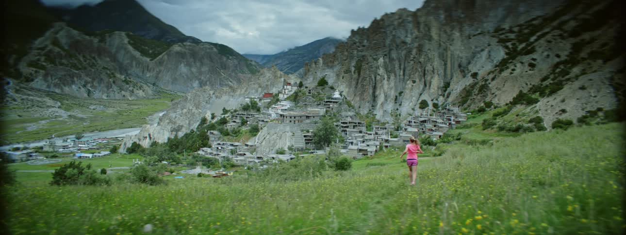 Woman jogging down a grassy field near mountains towards an old stone city