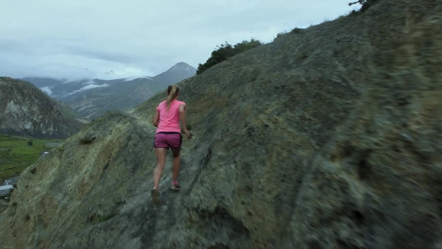 A woman runs along a narrow mountain trail near a rural neighborhood