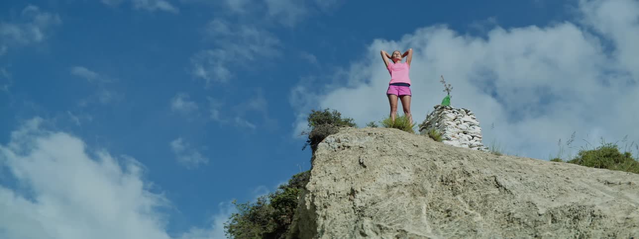 A woman stands near the edge of a cliff in the day and stretches her arms behind her head