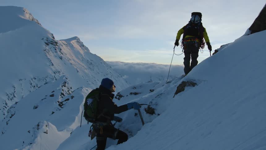 Two mountain climbers climb up the snow covered slopes of a mountain at evening