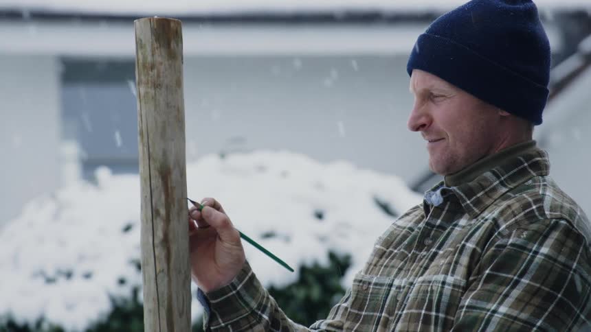 A man smiling and using a pencil to make marks on a wood post while it snows