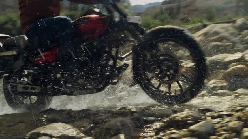 Motorcyclist rides motorcycle along rocky stream spraying water into the air