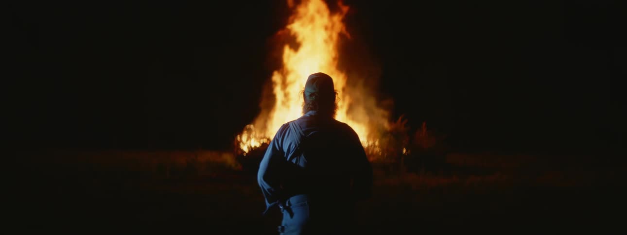 A man is standing outside in the darkness watching a huge bonfire burn as flames reach high into the air
