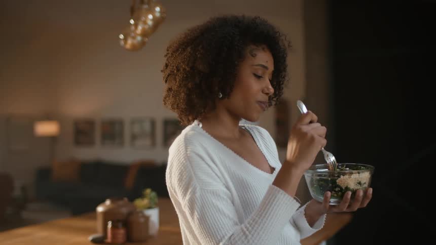 Young woman eats a bowl of salad reclined on the kitchen countertop at home