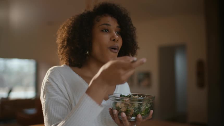 A woman is standing in a room holding a fork and a bowl as she smiles and eats a salad