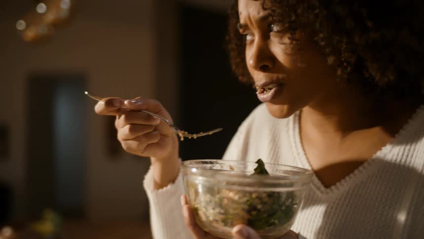 A woman stands in her kitchen eats a bite of salad and satisfactorily nods her head
