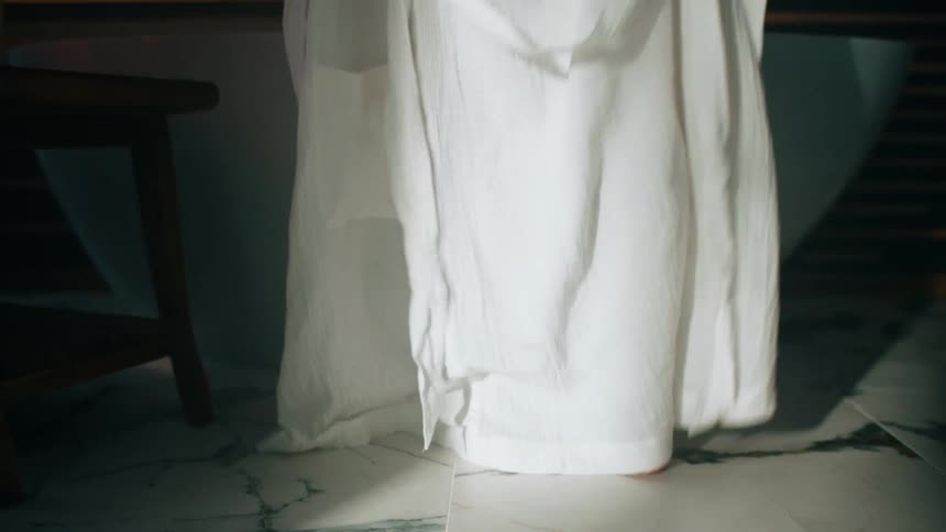 Close up of the legs of a woman dropping her white bathrobe on the bathroom floor before entering the bathtub
